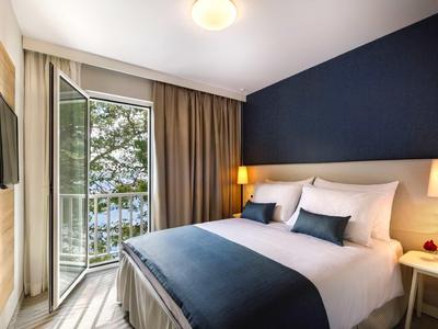 Chambre d'hôtel lumineuse avec lit double, tables de chevet et balcon donnant sur la verdure.