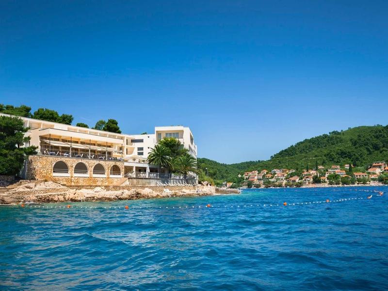 Hotel building on rocky shore with clear blue sea and forested hills in the background.