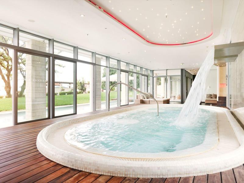 Moderní vnitřní bazén s vodopádem a velkými skleněnými okny s výhledem na zahradu.