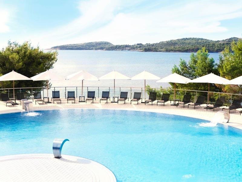 Kruhový hotelový bazén s lehátky a slunečníky s výhledem na moře v pozadí.