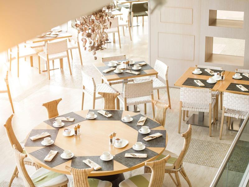 Světlá snídaňová místnost s kulatými a čtvercovými stolem, prostřenými šálky na kávu a talíři.