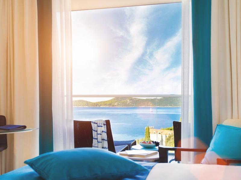 Výhled z hotelového pokoje na terasu s židlemi a moře pod jasnou oblohou.