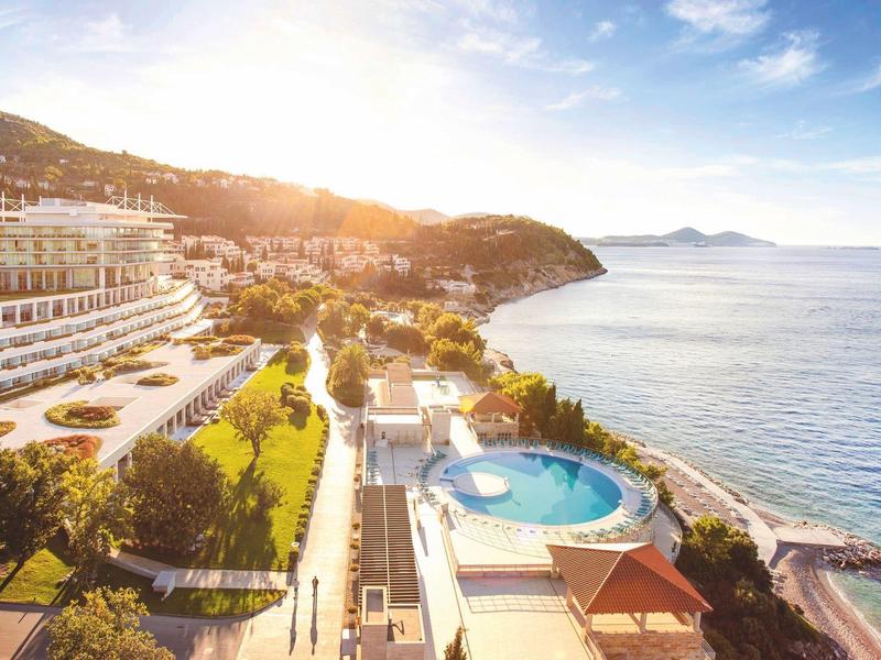 Luxusní hotel s bazénem u moře, obklopený zelenými kopci při západu slunce.