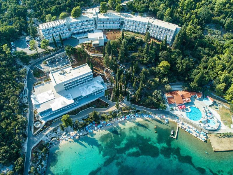 Aerial view of a hotel with pools, surrounded by forest and clear water.