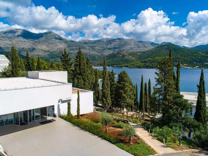 Modern hotel building with terrace by a lake, surrounded by mountains and trees under blue sky.