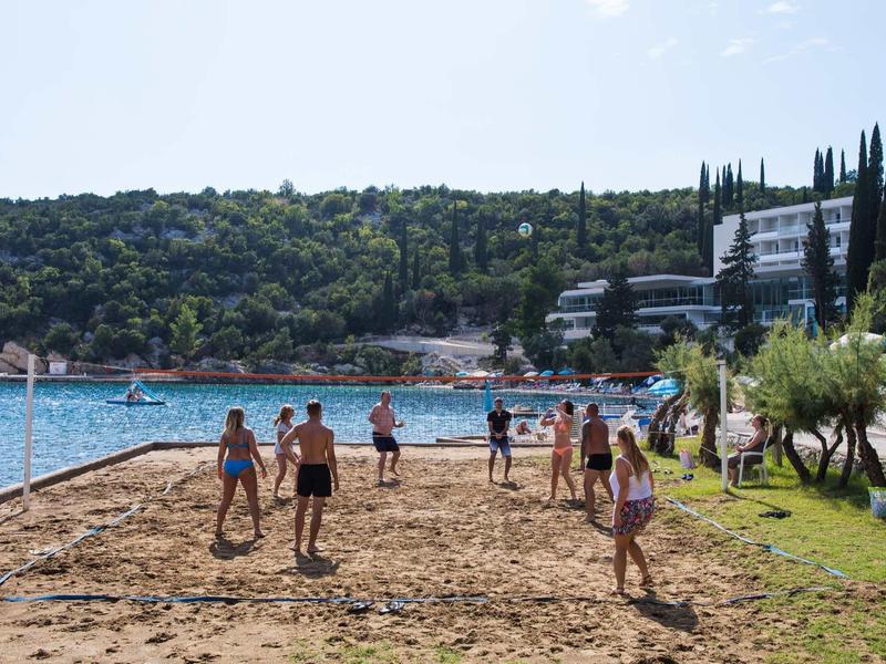 People playing volleyball on a sandy court by the water near a hotel with green hills in the background.