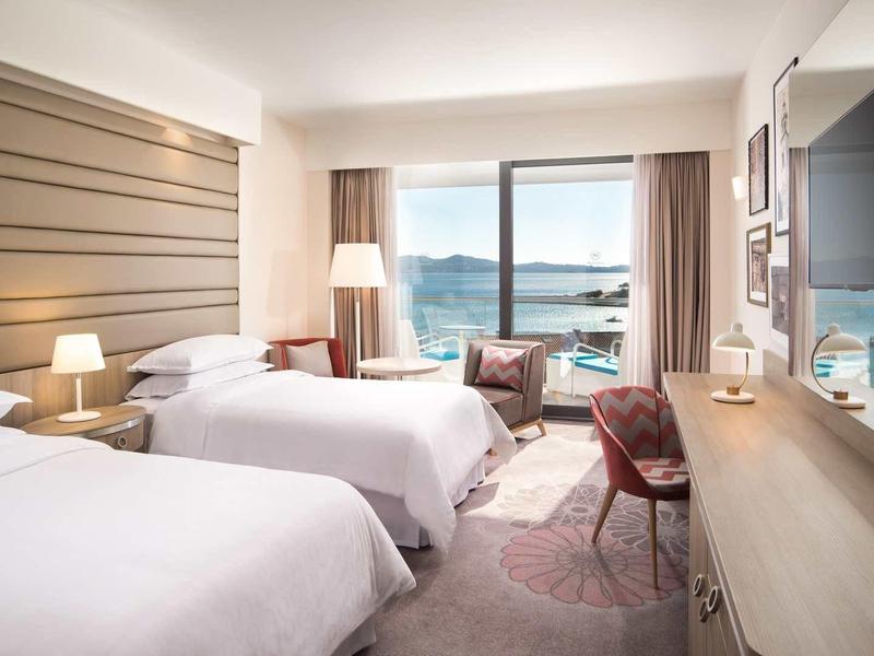 Helles Hotelzimmer mit zwei Betten, Schreibtisch, Stühlen und Meerblick durch große Fenster.