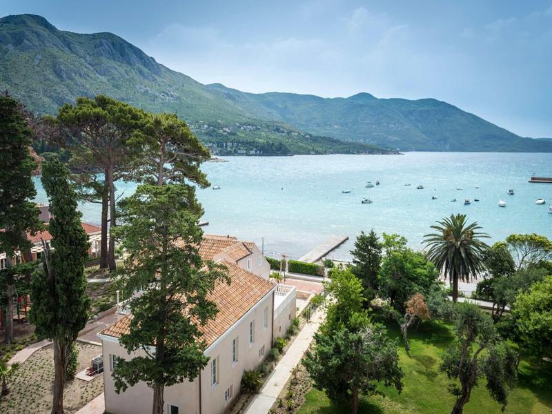Küste mit blauer Bucht, grünen Bäumen, Palmen und Bergen im Hintergrund unter klarem Himmel.