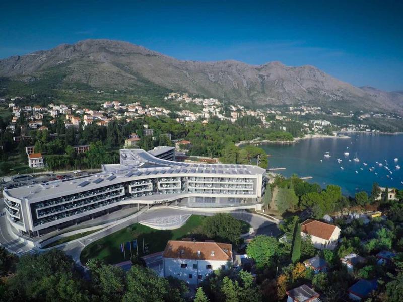 Modernes, geschwungenes Hotel an Küste mit Bergen, blauem Himmel und Segelbooten im Wasser.