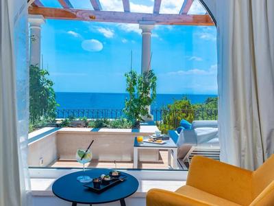 Vista da una camera con poltrona gialla e tavolo blu su una terrazza e il mare.