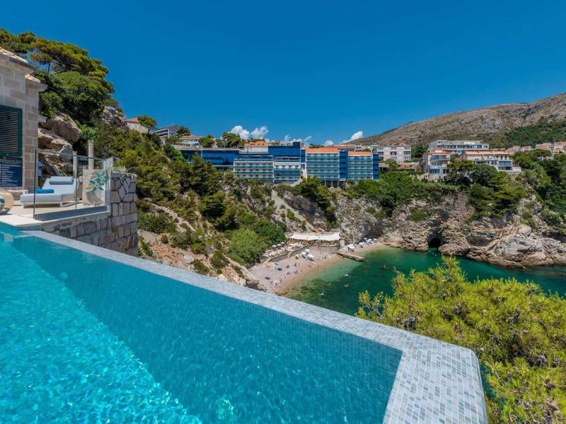 Piscina a sfioro con vista sulla costa, acque verdi e hotel sotto un cielo azzurro limpido.