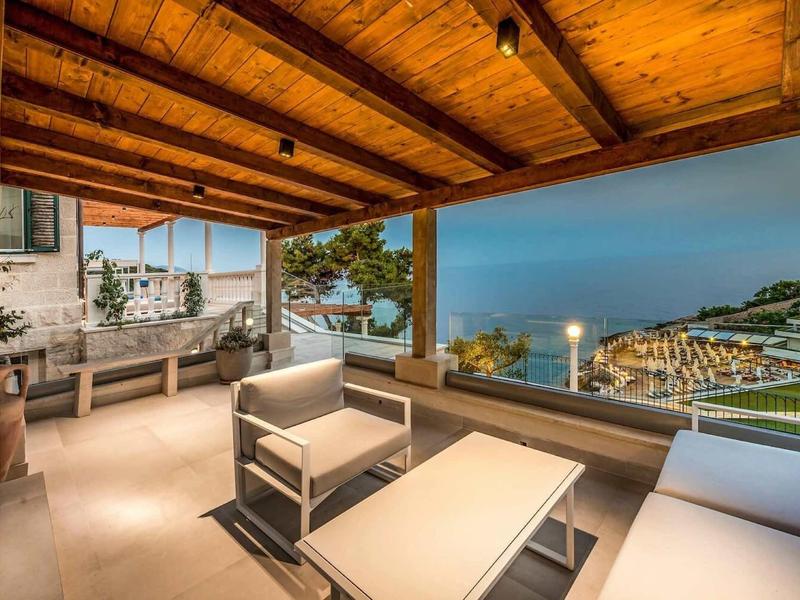 Terrazza coperta con zona soggiorno moderna e vista sui giardini dell'hotel illuminati e sul mare al crepuscolo.