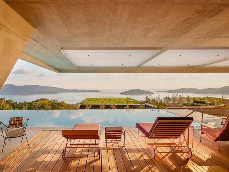 Moderner Poolbereich mit Holzdeck, Liegestühlen und Blick auf Meer und Inseln bei klarem Himmel.