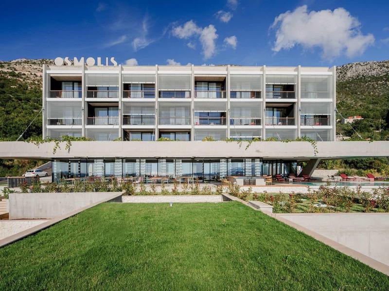 Modernes weißes Hotelgebäude mit großen Fenstern, Balkonreihen, grünem Rasen und Bergen im Hintergrund.