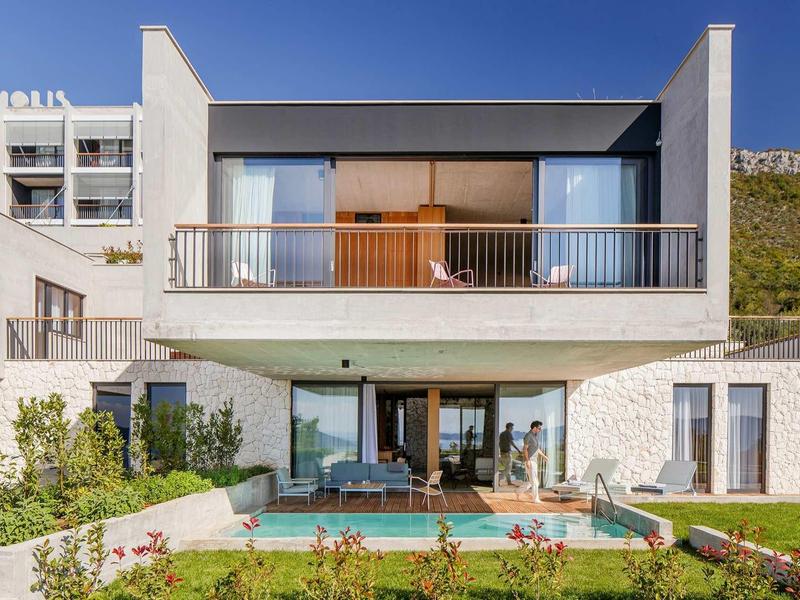 Modernes zweistöckiges Haus mit großer Glasfront, Balkon, Garten und kleinem Pool bei sonnigem Himmel.