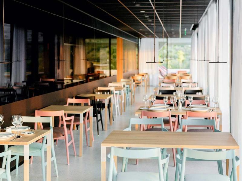 Helles modernes Restaurant mit Holztischen, bunten Stühlen und großen Fenstern an der rechten Seite.