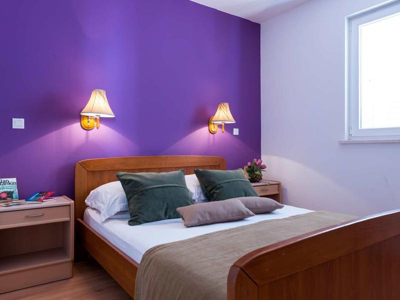 Schlafzimmer mit Holzbett, lila Wand, zwei grünen Kissen und Nachttisch mit Lampe und Büchern.