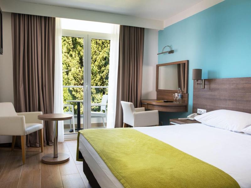 Camera d'hotel luminosa con letto grande, due sedie e vista balcone con alberi verdi.