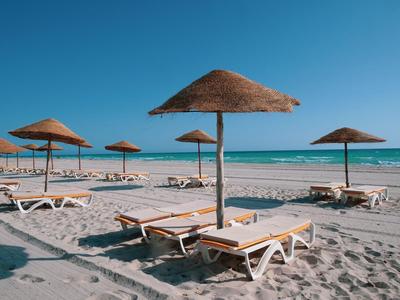 Strand mit Liegestühlen und Strohhutsonnenschirmen unter klarem blauem Himmel.