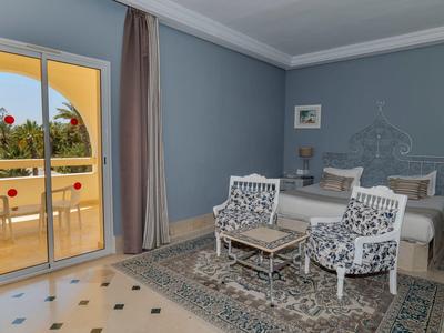 Chambre d'hôtel lumineuse avec lit double, deux fauteuils à motifs et balcon surplombant la verdure.