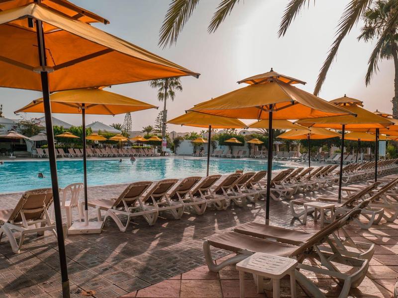 Rangées de chaises longues sous des parasols orange à côté d'une piscine avec des palmiers.