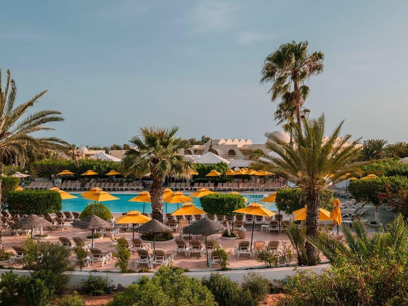Vue d'une piscine d'hôtel avec parasols et palmiers sous un ciel dégagé.
