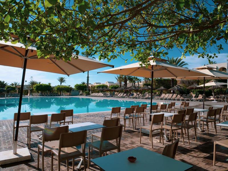 Terrasse d'hôtel avec chaises, tables et parasols près d'une piscine sous un ciel bleu.