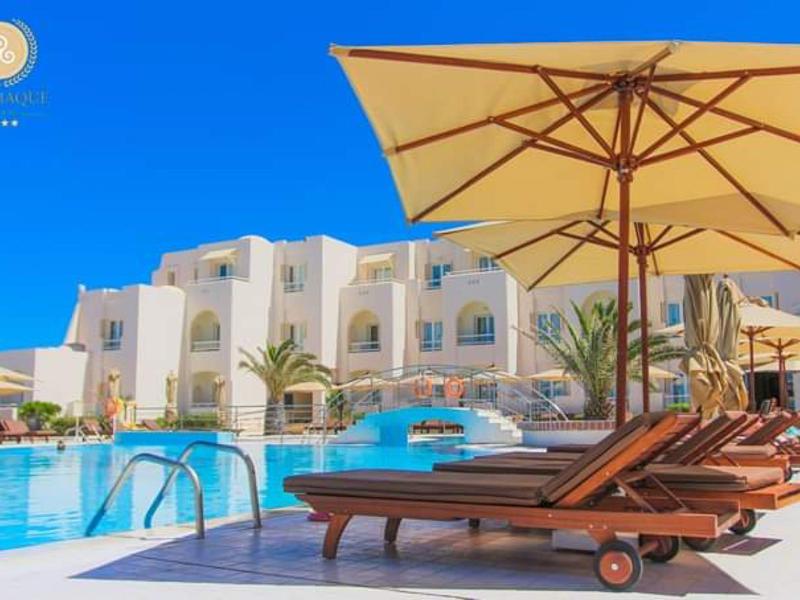 Zonovergoten resortzwembad met ligstoelen en parasols tegen een blauwe lucht en witte gebouwen.