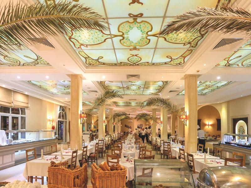 Ruime eetzaal van hotel met decoratief plafond, palmbomen en netjes gerangschikte tafels.