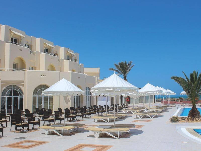 Zwembadterras van hotel met witte ligstoelen, parasols en een heldere blauwe lucht.