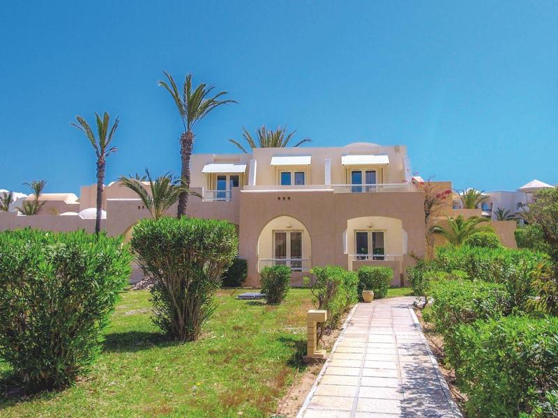 Pad leidt naar beige gebouwen omgeven door groen en palmbomen onder een heldere blauwe lucht