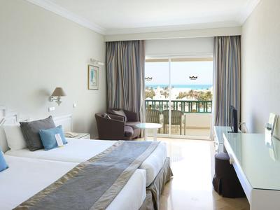 Helles Hotelzimmer mit zwei Betten, Balkon und Blick auf Gebäude.