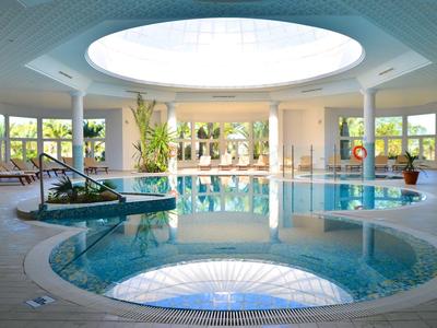 Heller Hallenbadbereich mit runder Deckenluke, Pflanzen und Loungemöbeln am Beckenrand.