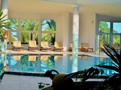 Heller Innenpool mit Liegestühlen, großen Fenstern und grünen Pflanzen im Hintergrund.