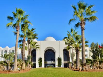 Modernes weißes Hotelgebäude mit Arkaden, umgeben von Palmen und grünem Rasen unter blauem Himmel