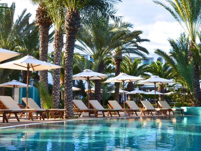 Großer Pool mit klar blauem Wasser, umgeben von Palmen und Sonnenliegen mit weißen Schirmen.