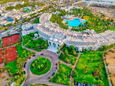 Großes, halbrundes Gebäude mit Pool, Tennisplätzen und viel Grün in sonniger Landschaft.
