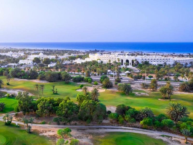 Grünes Golfresort mit Bäumen und Wegen, dahinter weiße Gebäude und blauer Meereshorizont.