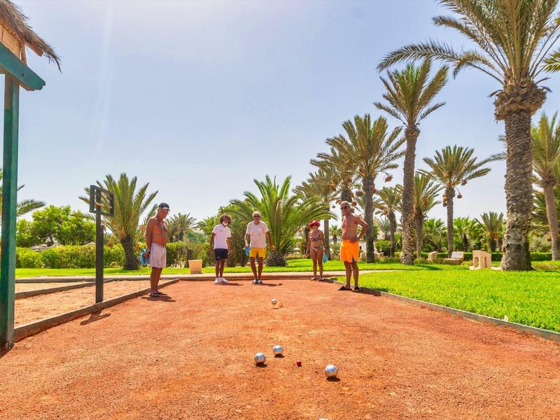 Menschen spielen Boule auf rotem Boden, umgeben von Palmen und grünem Gras bei sonnigem Wetter.