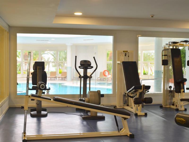 Fitnessraum mit Kraftgeräten, Laufband, Ergometer und großflächigen Fenstern mit Poolblick.