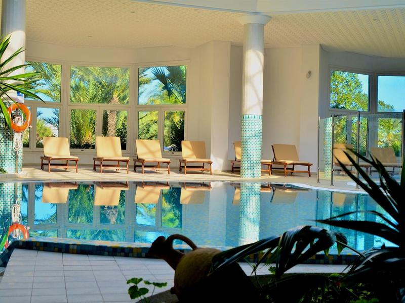 Heller Innenpool mit Liegestühlen, großen Fenstern und grünen Pflanzen im Hintergrund.