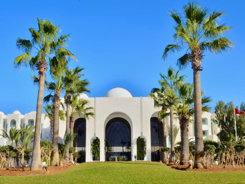 Weißes Gebäude mit Bögen und Palmen vor blauem Himmel auf gepflegtem Grün.