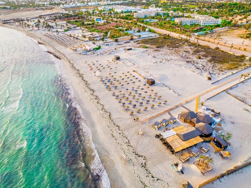 Ein langer Sandstrand mit Sonnenschirmen, Strandbars und klarem, grünlich-blauem Wasser.