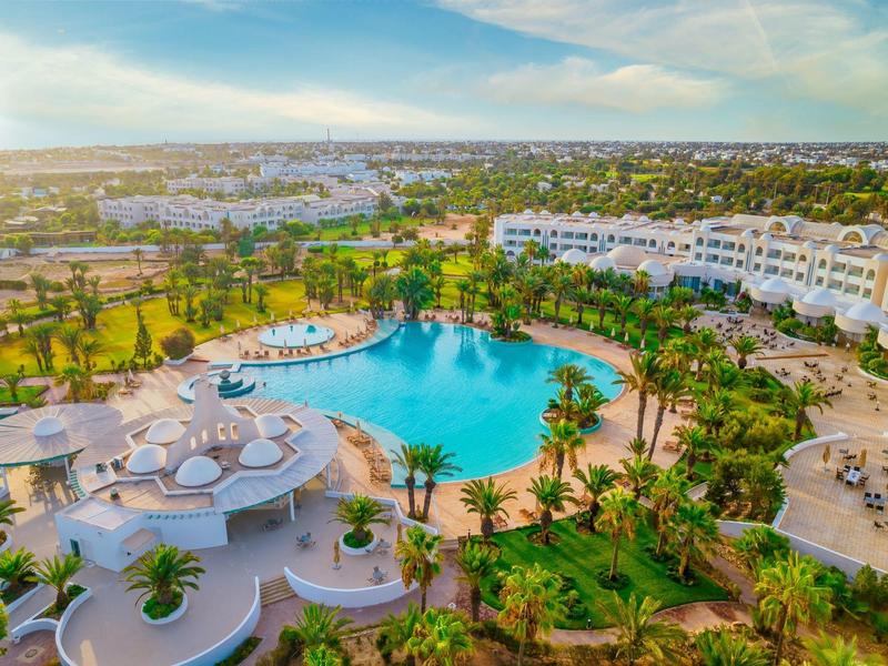 Große Hotelanlage mit rundem Pool, Palmen, grünen Rasenflächen und weißen Gebäuden bei Sonnenuntergang.