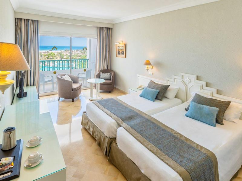 Helles Hotelzimmer mit zwei Betten, Sitzbereich, großem Fenster und Balkon mit Bergblick.