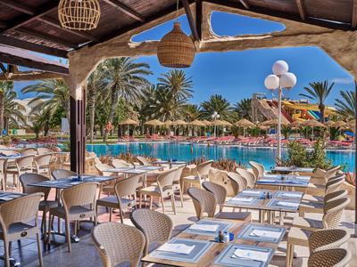 Restaurante al aire libre con mesas preparadas junto a una piscina y palmeras bajo un cielo azul