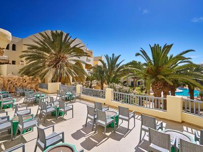 Außenbereich einer Terrasse mit Tischen und Stühlen neben einem Pool und Palmen unter blauem Himmel.