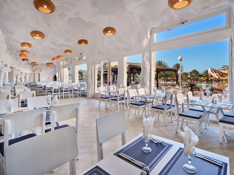 Restaurante elegante con muebles blancos modernos y vistas a una piscina y palmeras.