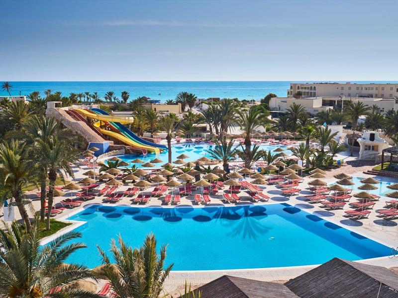 Gran piscina con toboganes, muchas tumbonas y palmeras en un complejo hotelero junto al mar.