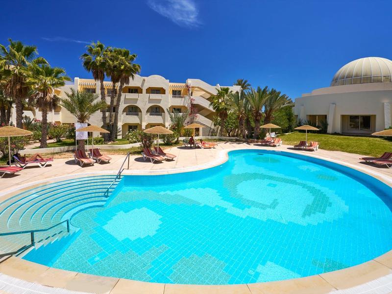 Piscina exterior redonda con tumbonas y palmeras frente a un hotel bajo un cielo despejado.